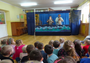 Scenografia teatralna - morze, dwóch aktorów z lalkami - rybakiem i żoną rybaka. Dzieci oglądające przedstawienie.